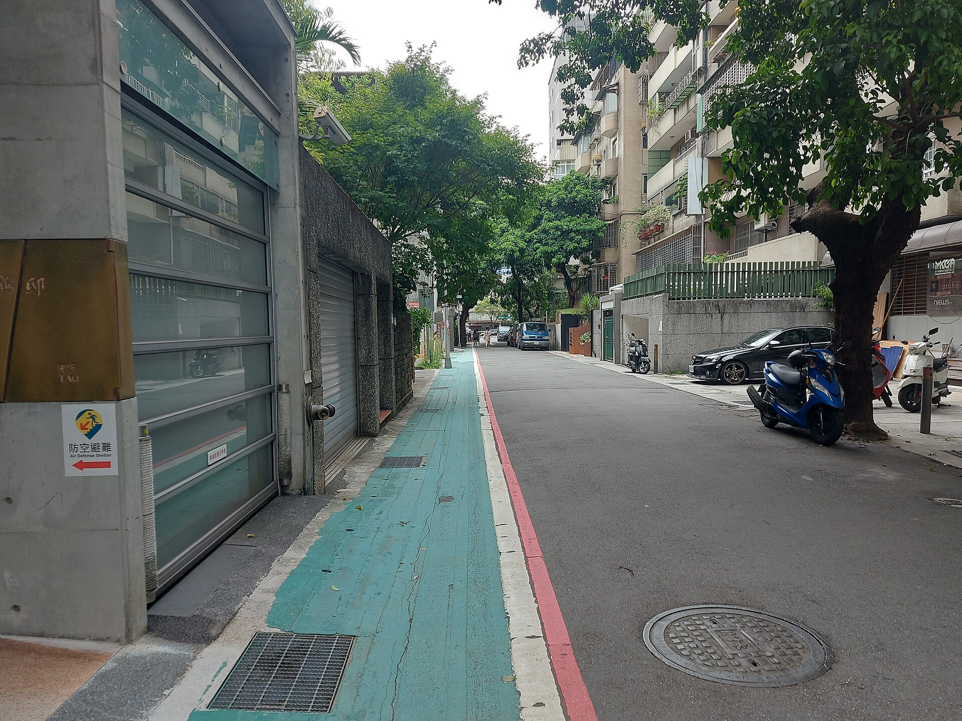 台北市内の並木道沿いの住宅街の路地、路肩に乗用車とスクーターが並び、奥に歩行者が小さく見える