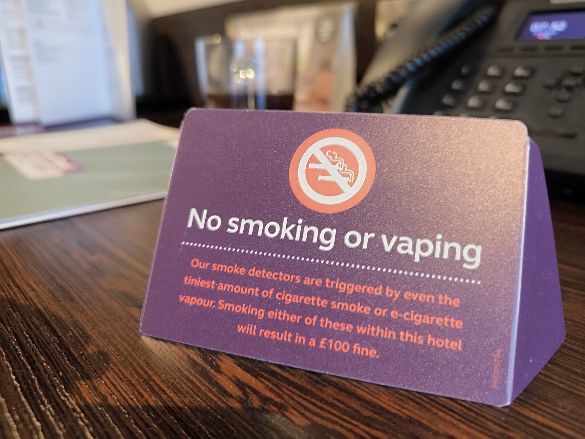 ホテルの机に置かれた紫色の「No smoking or vaping（喫煙・電子タバコ使用禁止）」の卓上サイン、禁煙マークと罰金の注意書きが書かれている
