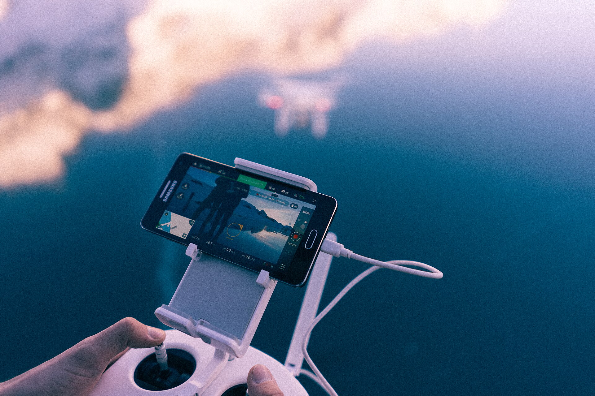 手に持ったコントローラーの画面を見ながら、曇り空を背景にぼんやりと浮かぶドローンを操縦している様子