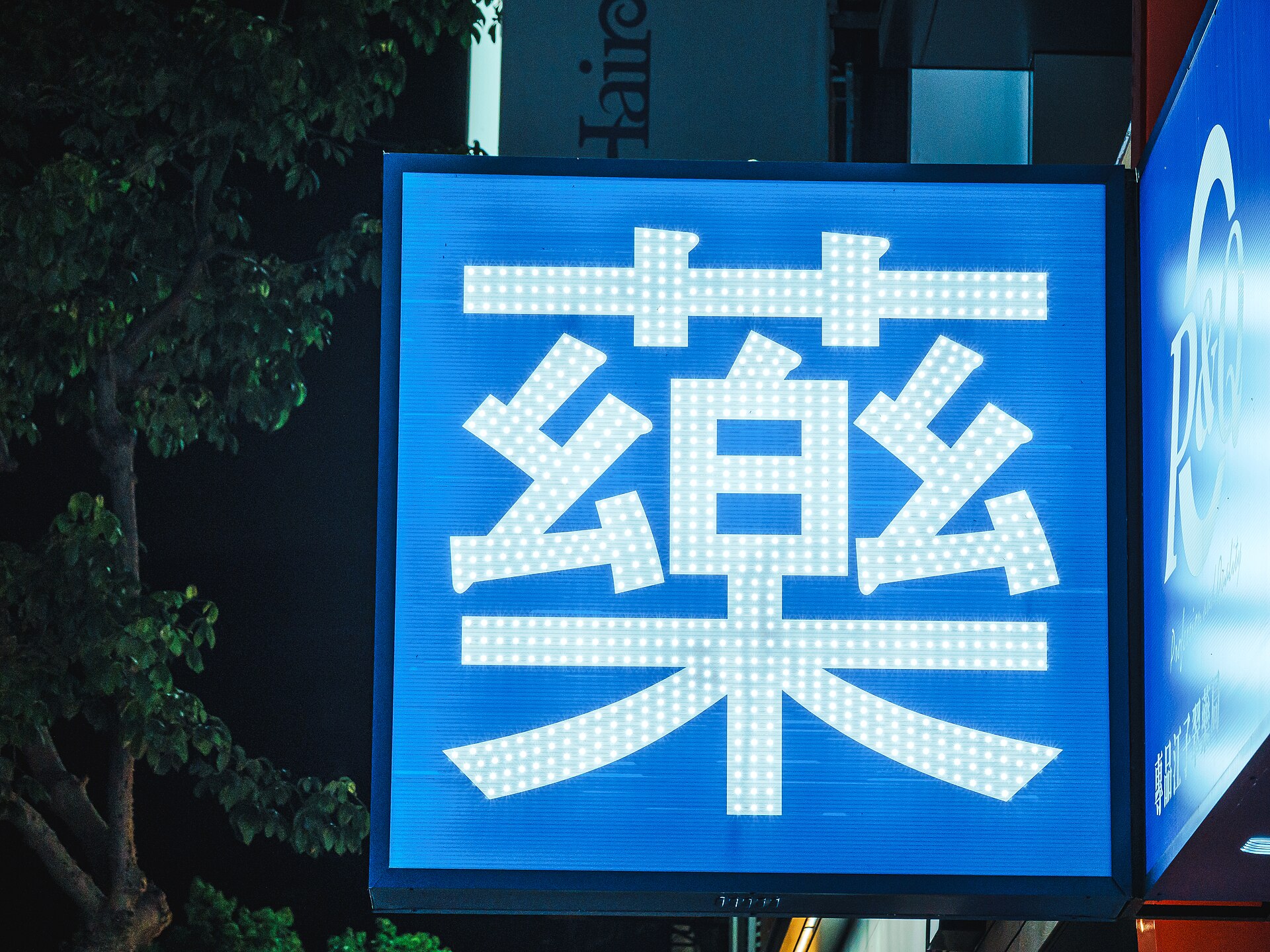夜の薬局の看板に灯る「薬」の文字のLEDイルミネーション、青い背景に白く浮かび上がっている